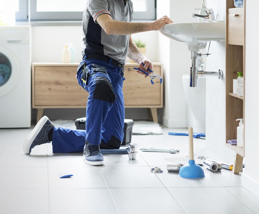Klempner kniet an einem Waschbecken, auf dem Boden liegen Werkzeuge